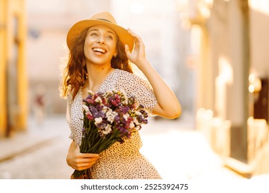 A joyful young woman on the cobblestone street, wearing a summer dress and a sun hat, holding a fresh bouquet of colorful flowers. Fashion style. - Powered by Shutterstock