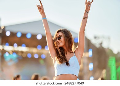 Joyful young woman raises hands in peace sign at beach music fest. Sunset glows, she dances, enjoys freedom, summer vibes. Sunglasses, crop top fashion, festival atmosphere, lively crowd.
