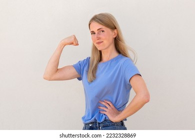 Joyful young woman showing her arm muscles. Caucasian female model with blonde hair and blue eyes in blue T-shirt smiling and demonstrating her bicep. Fitness, strength concept