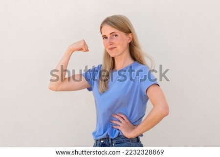 Joyful young woman showing her arm muscles. Caucasian female model with blonde hair and blue eyes in blue T-shirt smiling and demonstrating her bicep. Fitness, strength concept