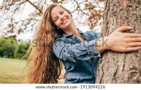 A joyful young woman smiling, wearing a blue denim shirt embraces a tree, posing on nature background. Happy female enjoying the time in the park. Mental health concept