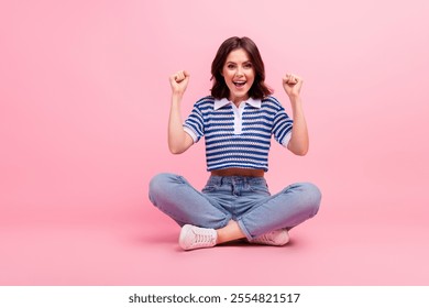 Joyful young woman in stylish casual attire showing excitement while sitting cross-legged against a pink backdrop - Powered by Shutterstock