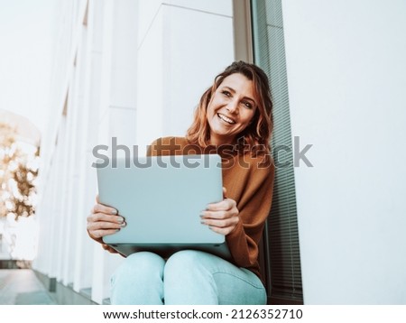 Joyful young woman with a vivacious smile seated on a windowsill of a commercial building in town with her laptop computer lookingup with a beaming smile