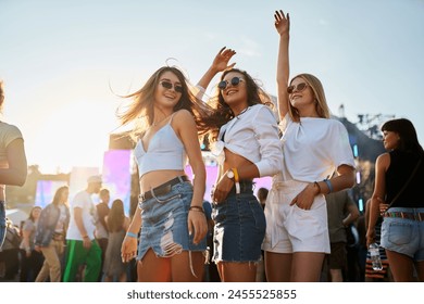Joyful young women dance at summer beach festival, friends enjoy live music, sunset vibes. Group in casual wear party, have fun in crowd near stage, sunny day. Female fans celebrate, outdoor event.