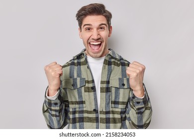Joyous adult man yells yes exclaims loudly celebrates something wears casual checkered shirt feels like winner isolated over grey studio background. Enthusiastic guy looks encouraged and confident - Shutterstock ID 2158234053