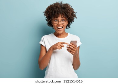 Joyous African American lady with big smile, points at screen of cell phone, rejoices having new app, enjoys online communication, being in high spirit, isolated over blue background. Technology - Shutterstock ID 1321514516