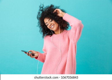 Joyous american woman in casual clothing dancing and listening to music with pleasure via white earphones isolated over blue background