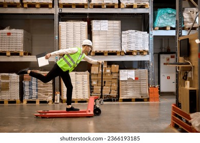 joyous caucasian male staff dressed in working clothes and helmet in warehouse having fun standing on pallet truck jack, driving among shelves, enjoy free time, leisure at work place - Powered by Shutterstock