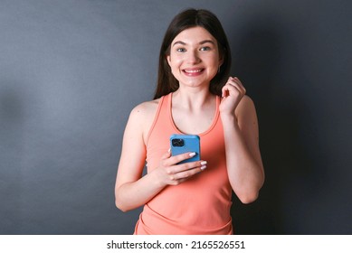 Joyous Caucasian woman wearing orange sportswear over studio grey wall poses with mobile phone device, types text message on modern smartphone, watches funny video during free time, enjoys internet