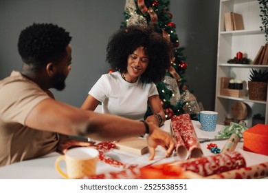 A joyous diverse couple sits at a table with wrapping paper and gifts, enveloped in Christmas cheer. The beautifully decorated tree enhances their festive holiday spirits. - Powered by Shutterstock