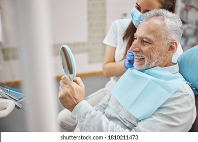 Joyous Elderly Man Sitting In A Dental Chair With A Doctor By His Side While Looking In The Mirror