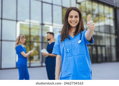 A joyous female nurse in blue scrubs displays a thumbs-up gesture in front of a medical facility with colleagues in the background engaged in conversation. - Powered by Shutterstock