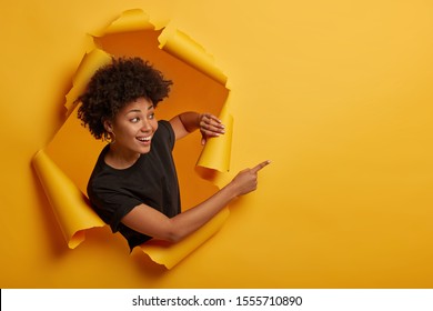 Joyous friendly looking smiling girl points aside with happy expression, toothy smile, pleased to show awesome advertisement, dressed casually says use copy space wisely gestures through ripped paper - Shutterstock ID 1555710890