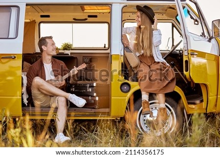 joyous hipster couple playing guitar ukulele sing a song in yellow van, in nature, field in countryside. traveler traveling together with vintage mini van in camping tent. camper vacation outdoors