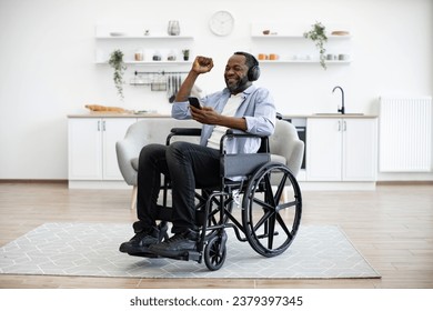 Joyous man in button-down shirt raising hand while looking at smartphone sitting in wheelchair in studio apartment. Cheerful african adult dancing along while hearing loud tunes in headphones. - Powered by Shutterstock