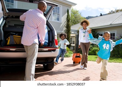 Joyous man loading luggage in back of a car as his daughter chases her brother with mom pulling a trolley bag outdoors.