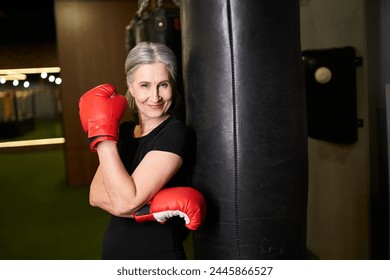 joyous mature woman in sportswear posing with boxing gloves near punching bag and smiling at camera - Powered by Shutterstock