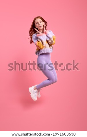 Joyous redhead woman in casual wear have fun alone in studio, posing at camera, jumping, holding hands on cheeks. Isolated pink studio background, portrait