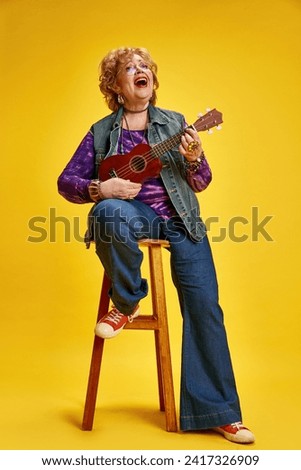 Joyous senior woman energetic playing ukulele seat on stool dressed casual denim and red shoes. Concept of empowerment of older adults, active seniors in modern life, fashion and style.