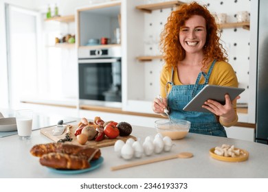 Joyous smiling young adult redhead woman looking at camera while searching food recipes on digital tablet in the kitchen, watching an online cooking class . - Powered by Shutterstock