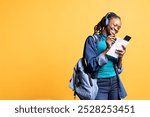 Joyous student taking notes for classes, transcribing info from smartphone screen and listening music, studio background. Young woman preparing for university, enjoying songs on headphones