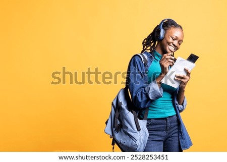 Joyous student taking notes for classes, transcribing info from smartphone screen and listening music, studio background. Young woman preparing for university, enjoying songs on headphones