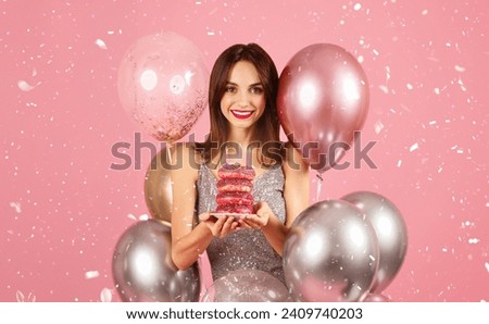 Joyous woman with a beaming smile celebrating with donuts topped with birthday candles, surrounded by a festive array of balloons amidst falling confetti on a pink background