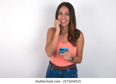 Joyous young beautiful caucasian woman wearing orange top over white background poses with mobile phone device, types text message on modern smartphone, watches funny video during free time.