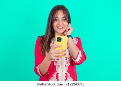 Joyous young beautiful moroccan woman wearing traditional caftan dress over green background poses with mobile phone device, types text message on modern smartphone, watches funny video .
