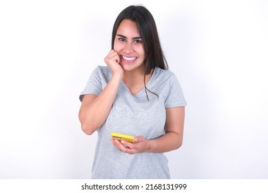 Joyous young caucasian woman wearing grey t-shirt over white background poses with mobile phone device, types text message on modern smartphone, watches funny video during free time, enjoys good 
