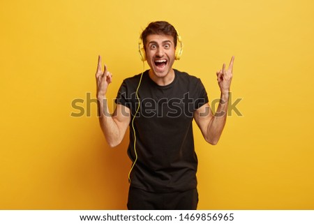 Joyous young hipster listens rock music in stereo headphones, shows heavy metal gesture, keeps mouth opened, dressed in black outfit, models against yellow background. Body language concept.