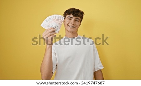 Joyous young hispanic teenager holding icelandic krona banknotes, radiating confidence while standing against a yellow isolated background