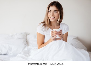 Joyous young lady looking to the side while relaxing in bed with a cup of tea - Powered by Shutterstock