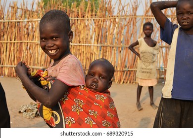 Juba, South Sudan, February 2017. Camp For Internally Displaced Persons (IDPs). A Little Smiling Girl Is Carrying Her Brother. Captured During Civil War.