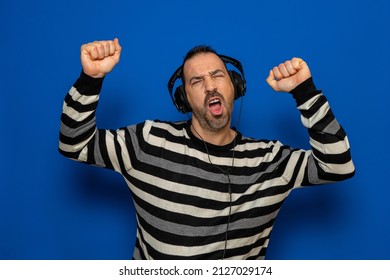 Jubilant 40 Year Old Hispanic Man Wearing Striped Pullover Listens To Music On Headphones And Dances Fun Isolated On Blue Background Studio Portrait