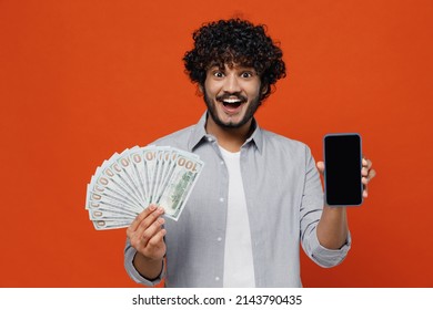 Jubilant exultant young bearded Indian man 20s years old wears blue shirt using mobile cell phone hold in hand fan of cash money in dollar banknotes isolated on plain orange background studio portrait