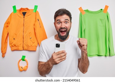 Jubilant happy bearded man clenches fist says yes uses mobile phone exclaims from joy wears white t shirt poses indoor with plastered jacket jumper and headphones. Yes finally I gained success