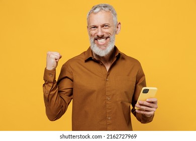 Jubilant happy elderly gray-haired bearded man 60s years old wears brown shirt look camera hold use mobile cell phone doing winner gesture celebrate isolated on plain yellow background studio portrait