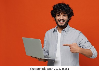 Jubilant happy exultant young bearded Indian man 20s years old wears blue shirt hold use work on laptop pc computer pointing index finger on screen isolated on plain orange background studio portrait