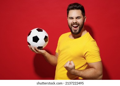 Jubilant happy young bearded man football fan in yellow t-shirt cheer up support favorite team hold soccer ball celebrate clenching fists say yes isolated on plain dark red background studio portrait