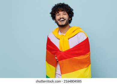 Jubilant vivid overjoyed fun excited young bearded Indian man 20s years old wears white t-shirt wrapped in striped colorful rainbow flag isolated on plain pastel light blue background studio portrait