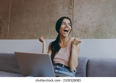 Jubilant young latin woman 20s in casual clothes sit on sofa hold use work on laptop pc computer celebrate clench fists look aside spend time in living room home own house be lost in reverie good day