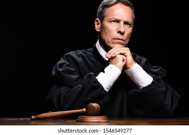 Judge In Judicial Robe Sitting At Table With Gavel Isolated On Black