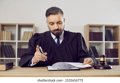 Judge working at his desk in court. Portrait of serious judge, lawyer or attorney dressed in robe gown uniform sitting at table with gavel hammer mallet, holding pen and reading judiciary law document