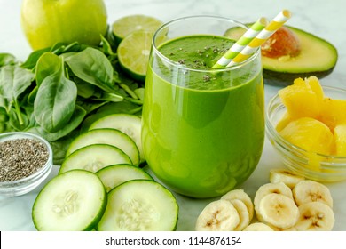 Juice Glass Filled With Green Kale And Spinach Smoothie With Green And Yellow Swirl Straws Surrounded By Ingredients Sitting On Marble Counter