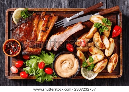 juicy grilled pork ribs with roasted potato wedges, fresh lettuce, tomatoes and mustard on wood rude board, horizontal view, flat lay, close-up