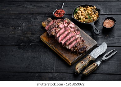 Juicy medium beef Rib Eye steak slices set, on wooden serving board, with white beans and rosemary in cast iron pan, with meat knife and fork, on black wooden table background with copy space for text