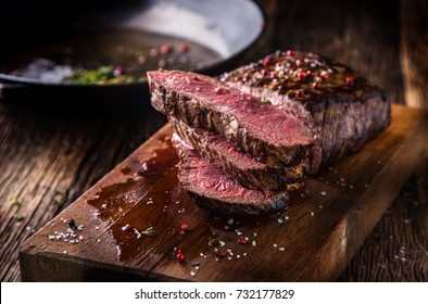 Juicy medium Beef Rib Eye steak slices in pan on wooden board with fork and knife herbs spices and salt.