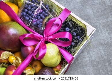 Juicy Ripe Fruits And Berries Are Beautifully Stacked In Wooden Box Tied With Pink Ribbon. Dark Background. Food Delivery, Gift Fruit Set. Concept Of Healthy Eating, Diet. Copy Space. Selective Focus.