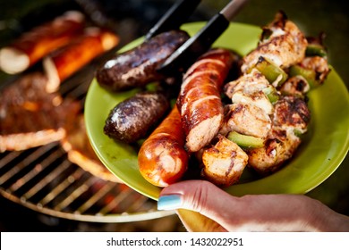 Juicy Sausages Straight From Grill On Green Plate Against The Background Of Hot Barbecue On Summer Day